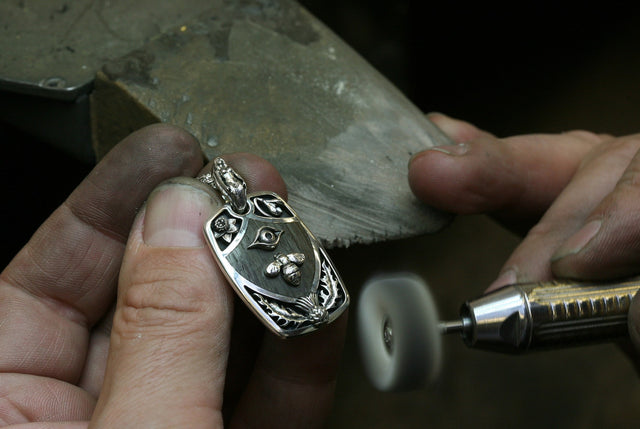A bespoke men's pendant being made in the Stephen Einhorn workshop. The pendant has been inlaid with thames wood and has thistles and a bee inlaid into it. The pendant is made from silver as are the inlays. We can see a polisher and the jewellers hands.
